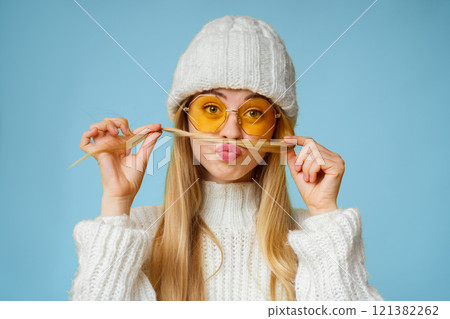 Smiling girl with sunglasses and winter hat making funny moustache out of her hair, yellow background 121382262