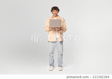 A young man wearing a beige shirt and blue jeans stands in a studio against a white background, smiling as he uses a laptop computer. A young man wearing a beige shirt and blue jeans stands in a studio against a white background, smiling as he uses a laptop computer. 121382264