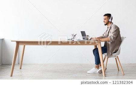 Cheerful Middle Eastern Freelancer Man Working On Pc Online At Home Listening To Music With Wireless Headphones, Making Research For New Project, Sitting At Desk Enjoying Remote Job, Profile Side View 121382304