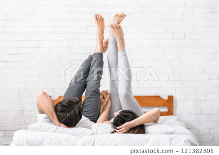 A couple lies in bed with their legs raised and hands clasped together. The woman is on her back, while the man is lying on his side. They are in a white bedroom with a brick wall behind them. A couple lies in bed with their legs raised and hands clasped together. The woman is on her back, while the man is lying on his side. They are in a white bedroom with a brick wall behind them. 121382305