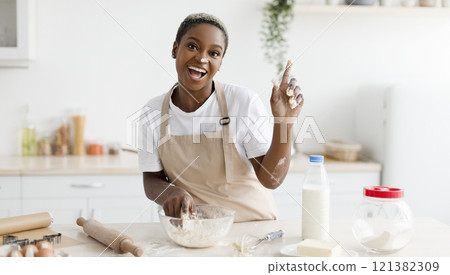 Laughing millennial african american woman in apron prepares dough for baking and raises finger up in light scandinavian kitchen interior. Got idea, cooking pizza, pie at home and facial expression 121382309