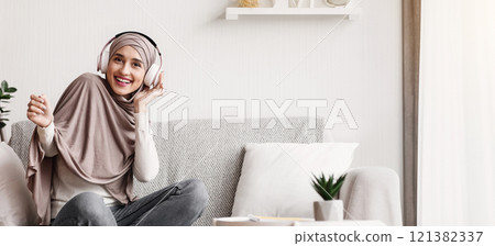 Cheerful arab girl sitting on sofa and listening music with wireless headphones, enjoying favorite song and smiling at camera Cheerful arab girl sitting on sofa and listening music with wireless headphones, enjoying favorite song and smiling at camera 121382337