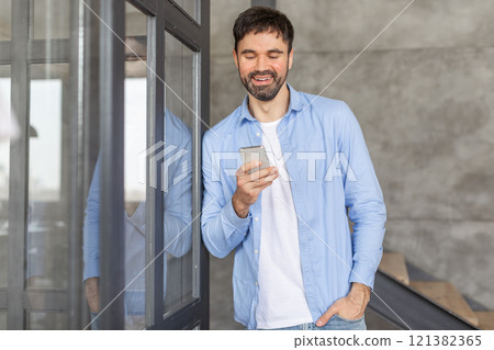 A smiling man in a light blue shirt and white t-shirt is standing by a glass door, looking down at his smartphone. 121382365