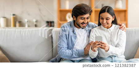 A young Indian couple smiles and shares a moment on a couch while looking at a smartphone. They appear relaxed and engaged, enjoying each other's company in a cozy living room. A young Indian couple smiles and shares a moment on a couch while looking at a smartphone. They appear relaxed and engaged, enjoying each other's company in a cozy living room. 121382387
