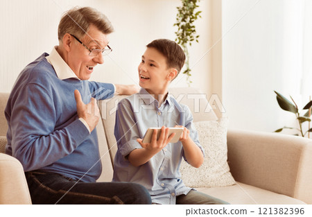 Smiling Boy Showing Grandfather How To Play Mobile Games On Smartphone Sitting On Sofa Indoor. Copy Space Smiling Boy Showing Grandfather How To Play Mobile Games On Smartphone Sitting On Sofa Indoor. Copy Space 121382396