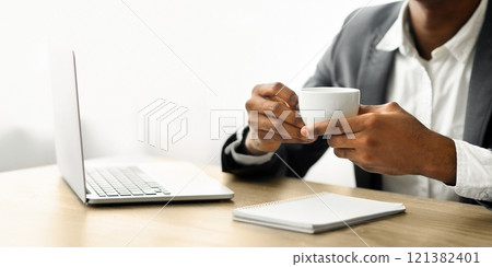 A businessman sits at a desk with a laptop and notebook, holding a white coffee cup. A businessman sits at a desk with a laptop and notebook, holding a white coffee cup. 121382401