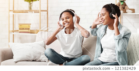 Two afro schoolgirls listening to music with wireless headphones and dancing at home, empty space Two afro schoolgirls listening to music with wireless headphones and dancing at home, empty space 121382429
