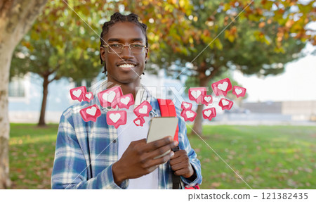 A young black man wearing a plaid shirt and glasses stands in a park with a backpack, smiling as he checks phone. Digital hearts and notifications float around him, suggesting social media activity. 121382435