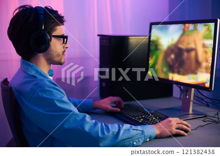 A young man wearing headphones is playing a computer game at a desk in his home office. He is focused on the screen and is using a keyboard and mouse to control the game. 121382438