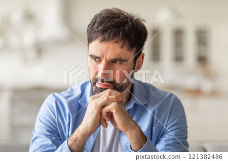A man with short brown hair and a beard is sitting indoors, wearing a blue button-down shirt. He is looking off to the side, resting his chin on his hands, and appears to be deep in thought. 121382486