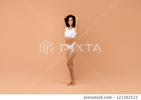 Black woman in white lingerie stands on a beige background, looking at the camera. She has dark, curly hair and is wearing a white bra and matching panties. Black woman in white lingerie stands on a beige background, looking at the camera. She has dark, curly hair and is wearing a white bra and matching panties. 121382513
