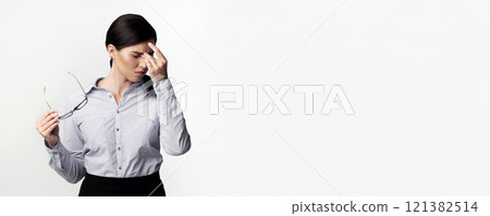 A woman in a light grey shirt and black skirt holds her glasses and rubs her forehead, looking stressed and suffering from a headache. A woman in a light grey shirt and black skirt holds her glasses and rubs her forehead, looking stressed and suffering from a headache. 121382514