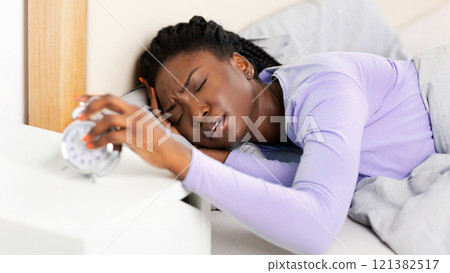 Sleepy Black Girl Turning Off The Alarm Clock Lying In Bed At Home In The Morning. Selective Focus Sleepy Black Girl Turning Off The Alarm Clock Lying In Bed At Home In The Morning. Selective Focus 121382517