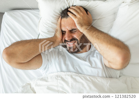 A man lies in bed, his face contorted in pain as he holds his head with both hands. He is likely suffering from a headache or migraine, his expression showing discomfort and distress, top view 121382520