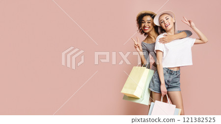 Two women, one in a striped shirt and a hat, and the other in a white shirt and denim shorts, hold shopping bags and smile at the camera. They are standing against a pink background. Two women, one in a striped shirt and a hat, and the other in a white shirt and denim shorts, hold shopping bags and smile at the camera. They are standing against a pink background. 121382525