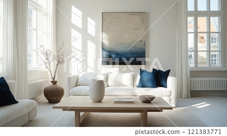 The spacious living room has a white sofa with blue pillows, a light wooden coffee table, and an abstract painting, all bathed in soft natural light from large windows. 121383771