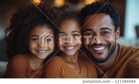 A father and his two daughters share a warm embrace, smiling broadly while enjoying a cozy indoor moment together. They are dressed in matching autumn colors that enhance the joyful atmosphere. 121384084