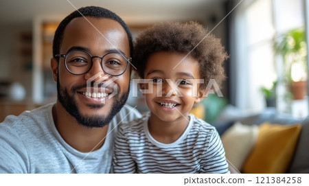 A father and his young son sit together in a warm living room, both smiling widely. They enjoy each others company, surrounded by soft furnishings and sunlight filtering through the windows. 121384258