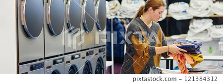 Website header shot of young woman putting pile of clean clothes into basket on folding table in self service laundry room, copy space Website header shot of young woman putting pile of clean clothes into basket on folding table in self service laundry room, copy space 121384312