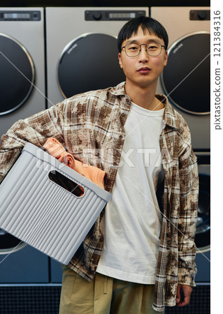 Vertical shot of young Asian man carrying basket with clothes preparing to load washing machine while confidently looking at camera in self service laundry Vertical shot of young Asian man carrying basket with clothes preparing to load washing machine while confidently looking at camera in self service laundry 121384316