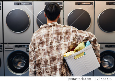 Rear view of young man dressed in plaid shirt holding basket of clothes ready for washing in tumbling machines in background in self service laundry, copy space Rear view of young man dressed in plaid shirt holding basket of clothes ready for washing in tumbling machines in background in self service laundry, copy space 121384320