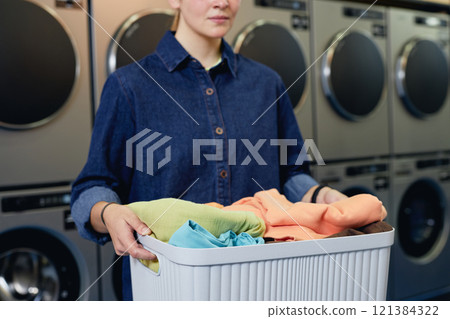 Cropped shot of girl holding basket with load of laundry preparing colored clothes for washing at self service laundromat, copy space Cropped shot of girl holding basket with load of laundry preparing colored clothes for washing at self service laundromat, copy space 121384322