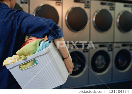 Back view of unrecognizable girl carrying basket with sorted clothes preparing colored garments for washing in self service laundry, copy space Back view of unrecognizable girl carrying basket with sorted clothes preparing colored garments for washing in self service laundry, copy space 121384324
