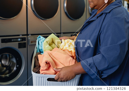 Cropped shot of senior African American woman carrying basket with sorted clothes preparing colored clothes for washing at modern self service laundry room, copy space 121384326