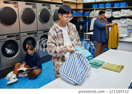 Medium shot of young Asian man packing clean clothes into bag on folding table checking results after washing garments in public laundry room, copy space 121384332