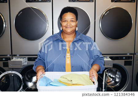 Portrait of happy African American female customer holding basket with clothes smiling at camera during daily domestic routine in self service laundry, camera flash, copy space 121384346