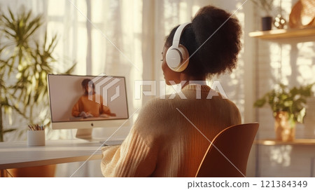A woman attentively participates in an online class from her cozy home workspace. She is wearing headphones while focusing on the computer screen, surrounded by plants and soft lighting. A woman attentively participates in an online class from her cozy home workspace. She is wearing headphones while focusing on the computer screen, surrounded by plants and soft lighting. 121384349