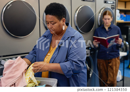 Medium shot of senior African American woman carefully sorting through colored clothes in basket on folding table while organizing daily laundry at coin operated laundromat, copy space Medium shot of senior African American woman carefully sorting through colored clothes in basket on folding table while organizing daily laundry at coin operated laundromat, copy space 121384350