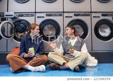 Young woman and Asian man engaged in lively conversation sitting on floor passing time, while washing machines tumbling with clothes in background in self service laundry 121384359