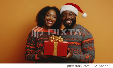 A joyful couple poses together, both wearing matching festive sweaters, while holding a red gift box wrapped with a golden bow. Their smiles radiate holiday cheer against a warm backdrop. A joyful couple poses together, both wearing matching festive sweaters, while holding a red gift box wrapped with a golden bow. Their smiles radiate holiday cheer against a warm backdrop. 121384368