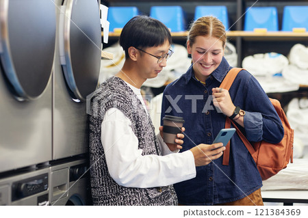 Medium shot of young Asian man showing video on phone to cheerful female friend while washing clothes and waiting for result at self service laundry 121384369