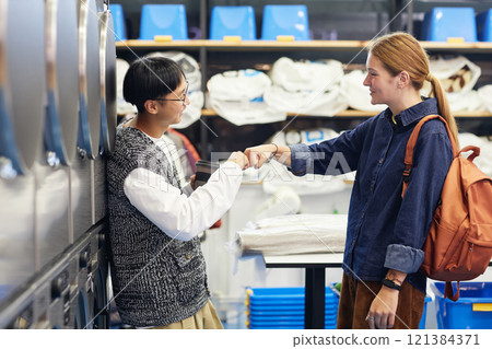 Side view of happy Asian man bumping fists with female friend meeting at self service laundry while washing clothes and waiting for result 121384371