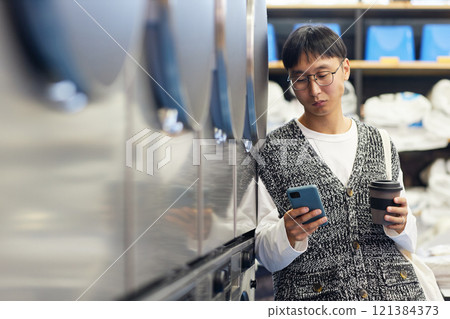 Medium shot of young Asian man with cup of coffee in hand text messaging on phone while waiting in coin laundromat standing next to line of washing machines, copy space 121384373