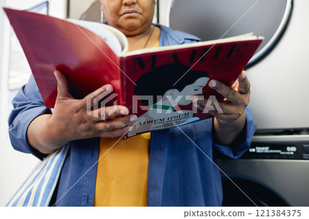 Cropped shot of senior African American woman reading fashion magazine while passing time in self service laundry room waiting for end of washing, copy space 121384375