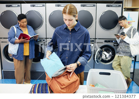 Medium shot of girl in casual denim shirt carefully packing clean clothes into backpack on folding table checking results of washing with eco friendly detergent in public laundry, copy space 121384377