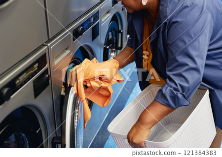 Cropped shot of senior African American woman loading colored clothes from basket into washing machine while doing daily laundry at modern self service laundromat 121384383