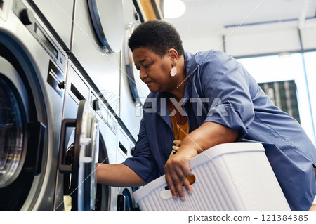 Medium shot of senior African American woman with laundry basket in hands loading clothes into washing machine at coin operated laundromat, household chores concept 121384385