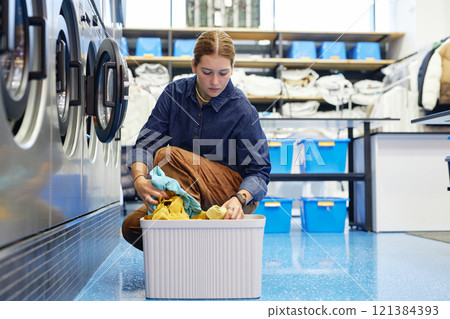 Young woman sorting laundry in basket loading clothes into washing machine while doing domestic chores at laundromat using cheap and convenient self service, copy space 121384393