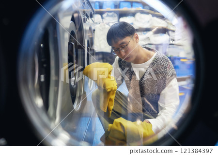 Shot through open washing machine of young Asian man unloading clean clothes while doing daily laundry at coin laundromat, household chores concept Shot through open washing machine of young Asian man unloading clean clothes while doing daily laundry at coin laundromat, household chores concept 121384397