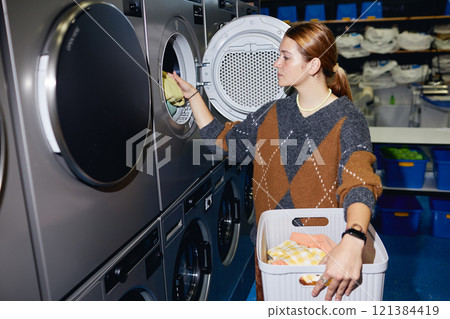Side view of young woman holding basket in hands loading washing machine with clothes in modern self service laundry, camera flash 121384419