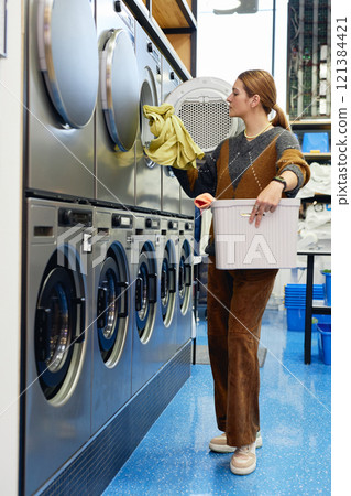 Vertical shot of young woman dressed in vintage outfit with basket in hands loading washing machine with clothes in contemporary self service laundry room 121384421
