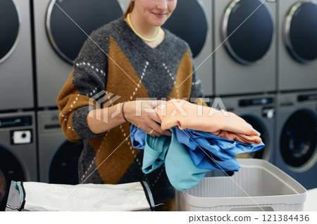 Cropped shot of young woman holding pile of colored clothes sorting delicate garment before loading into washing machine in self service laundry, copy space Cropped shot of young woman holding pile of colored clothes sorting delicate garment before loading into washing machine in self service laundry, copy space 121384436