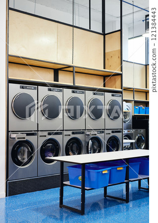 Interior of modern self service laundry featuring double row of professional washing machines and clothes dryers nest to folding table, copy space Interior of modern self service laundry featuring double row of professional washing machines and clothes dryers nest to folding table, copy space 121384443