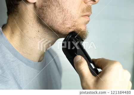 A man trims his beard with a trimmer. An electric razor trimming a beard. Beard care at home. A man trims his beard with a trimmer. An electric razor trimming a beard. Beard care at home. 121384480