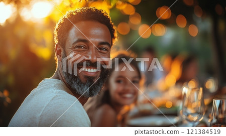 A joyful man gazes at the camera, sitting at a beautifully set table outdoors during sunset, surrounded by friends and warm lights. 121384519
