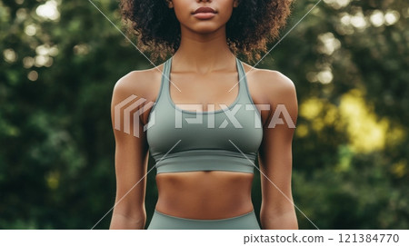 A young woman with curly hair stands tall in athletic gear, showcasing her fitness and determination in a sunlit outdoor setting, surrounded by greenery, during an afternoon workout session. 121384770
